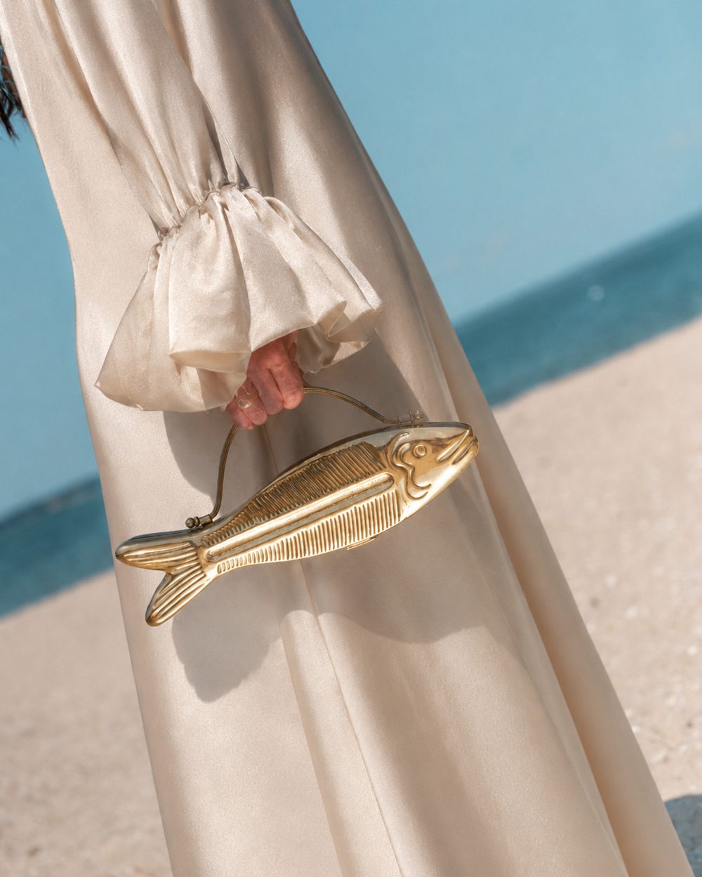 Azure Koi clutch carried by its chain handle, styled with a flowing cream dress on a beach